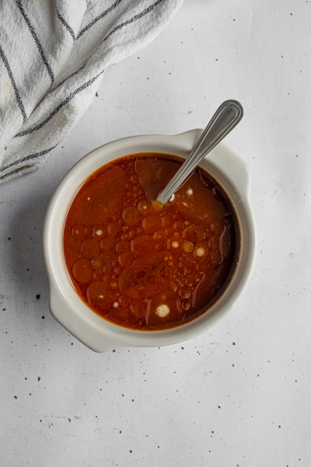 A small ceramic bowl with a spoon stirring a sesame stir fry sauce. The bowl is on a white counter with a white striped napkin.