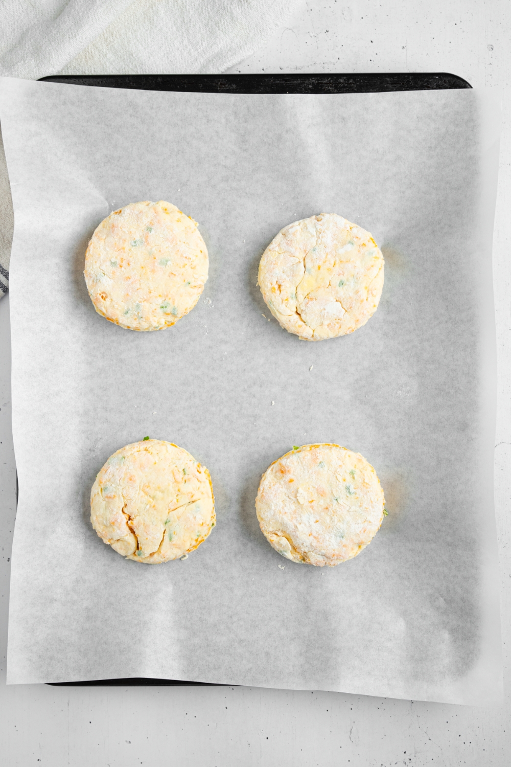 Four uncooked cheddar chive biscuits on a baking sheet lined with parchment paper. The sheet is on a white counter.
