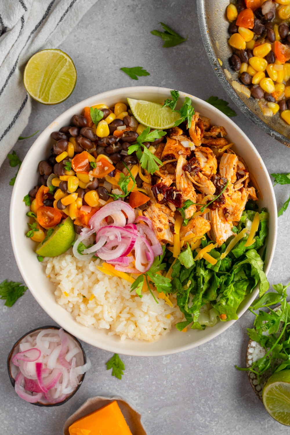 A white ceramic bowl with rotisserie chicken burrito bowl. The bowl includes white rice, rotisserie chicken, corn, beans, and pickled red onion garnished with fresh cilantro. The bowl is on a white counter with lime, pickled red onion, and a bowl of corn salsa.