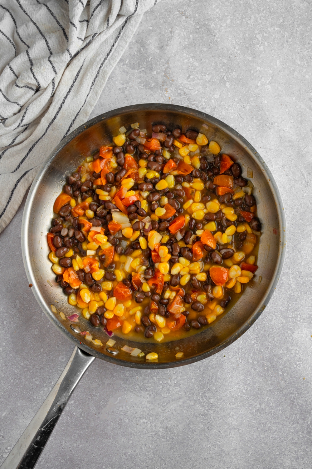 A skillet with corn, beans, and tomatoes cooking in oil. The skillet is on a white counter with a white striped napkin.