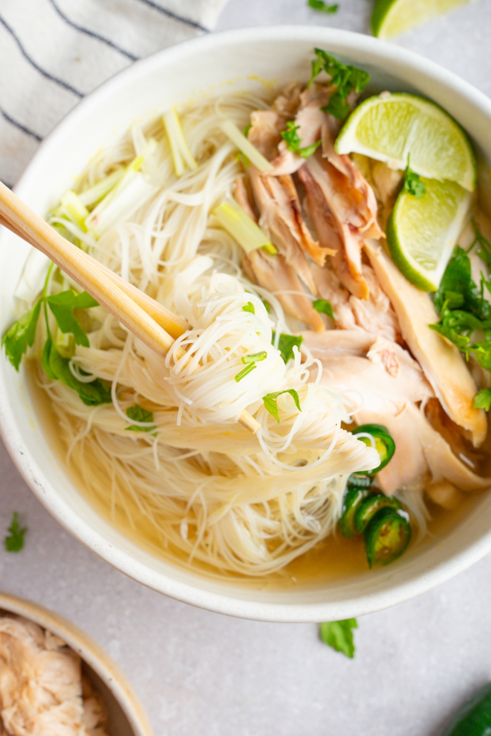 Rotisserie chicken pho served in a white bowl garnished with lime, fresh basil leaves, cilantro, bean sprouts, sliced jalapeno, and green onions. A pair of chopsticks is taking a bite of pho noodles. The bowl is on a white counter with a white striped napkin.