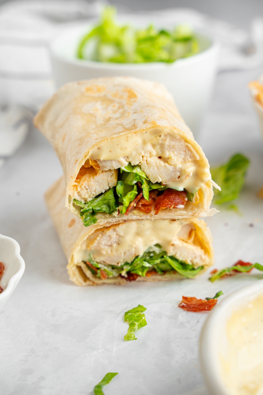 A rotisserie chicken caesar wrap sliced in half and stacked on one another. The wrap is on a white counter with a bowl of shredded lettuce.