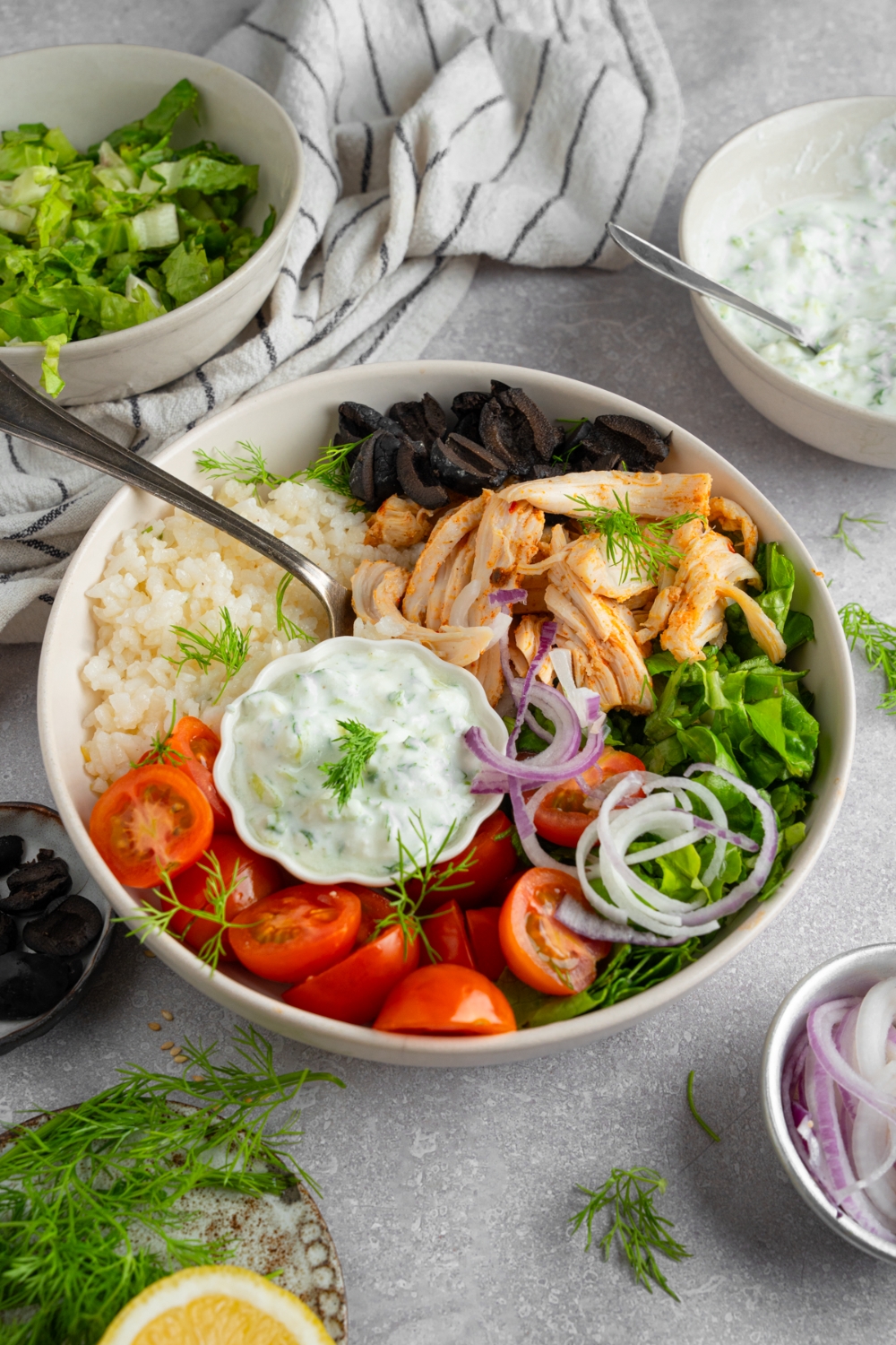 Rotisserie chicken greek bowl served in a white bowl with lettuce, rotisserie chicken, rice, black olives, tomatoes, red onion, dill, and a small bowl of tzatziki. There is a fork in the bowl. The bowl is on a white counter with a small bowl of lettuce and small bowl of tzatziki.