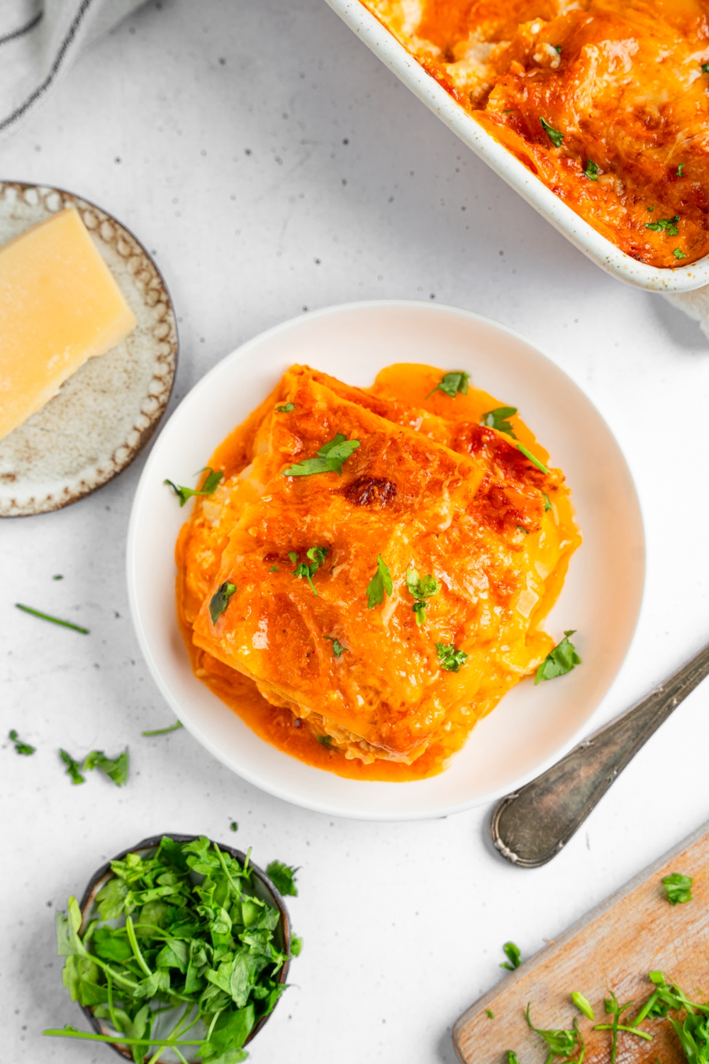 Rotisserie chicken lasagna served on a white plate garnished with fresh basil. The plate is on a white counter with a baking dish of lasagna and small plates of garnishes.
