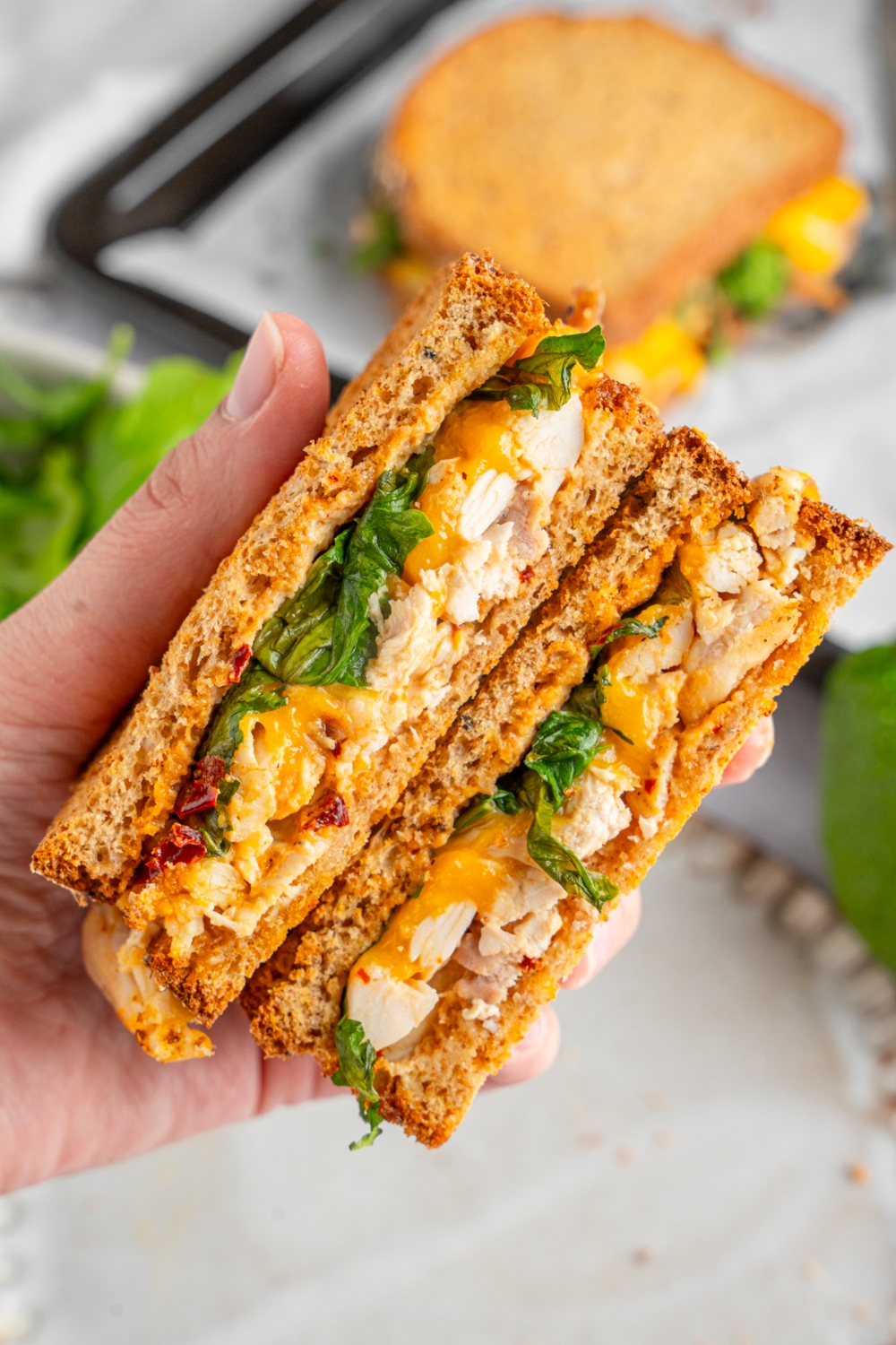 A hand holding a rotisserie chicken sandwich sliced in half. There is a ceramic plate and lined baking sheet with an additional sandwich blurred in the background.