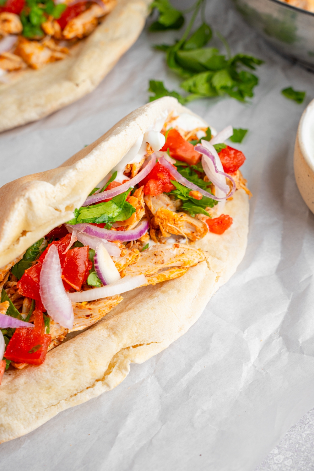 A folded chicken shawarma including a pita wrapped with rotisserie chicken, garlic sauce, red onion, tomatoes, and fresh parsley. The shawarma is on a piece of parchment paper with an additional pita and fresh parsley.