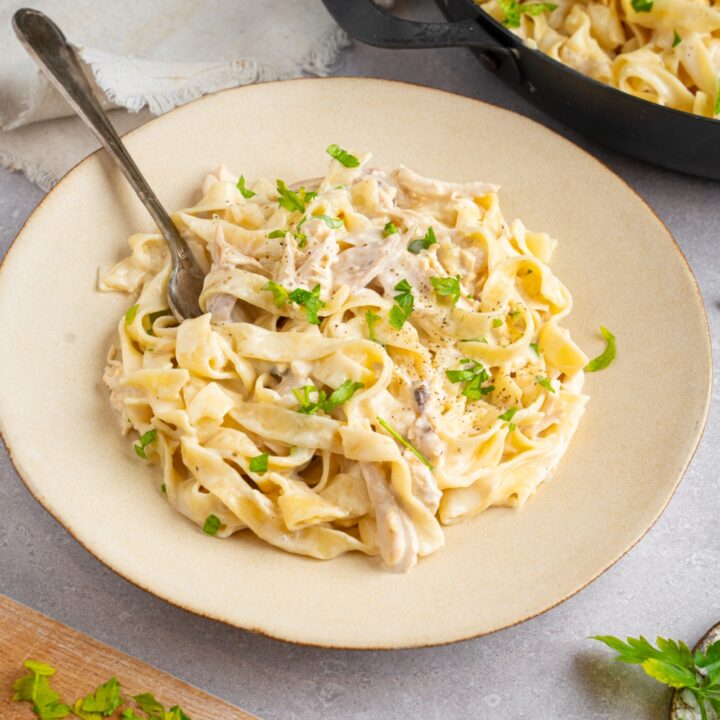 A white ceramic plate with rotisserie chicken fettuccine alfredo. There is a fork in the plate and the pasta is garnished with fresh parsley. The plate is on a white counter with a skillet of pasta and white cloth napkin.