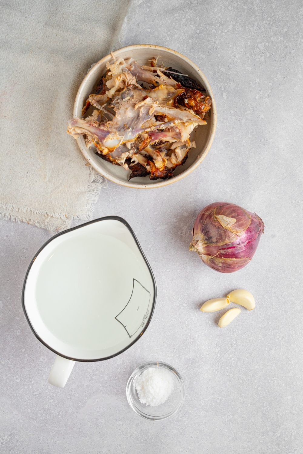 An overhead shot of several bowls in various sizes containing ingredients to make rotisserie chicken bone broth including water, rotisserie chicken, onion, garlic, and salt.