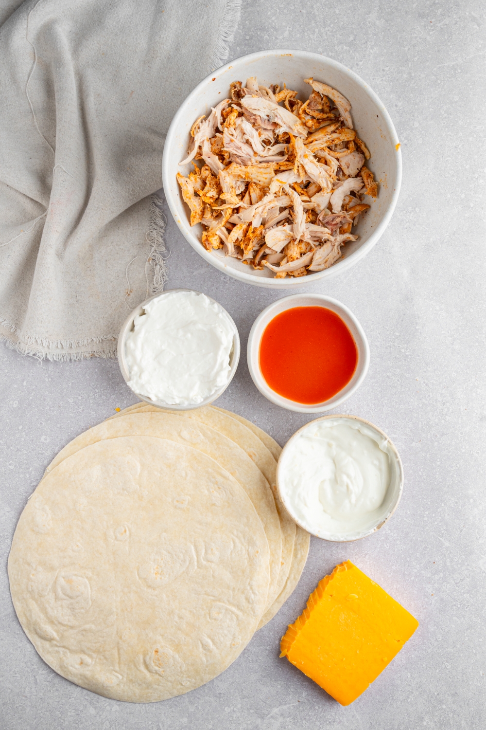 An overhead shot of several bowls in various sizes containing ingredients to make rotisserie chicken burritos including shredded rotisserie chicken, tortillas, cream cheese, greek yogurt, buffalo sauce, and cheddar cheese.