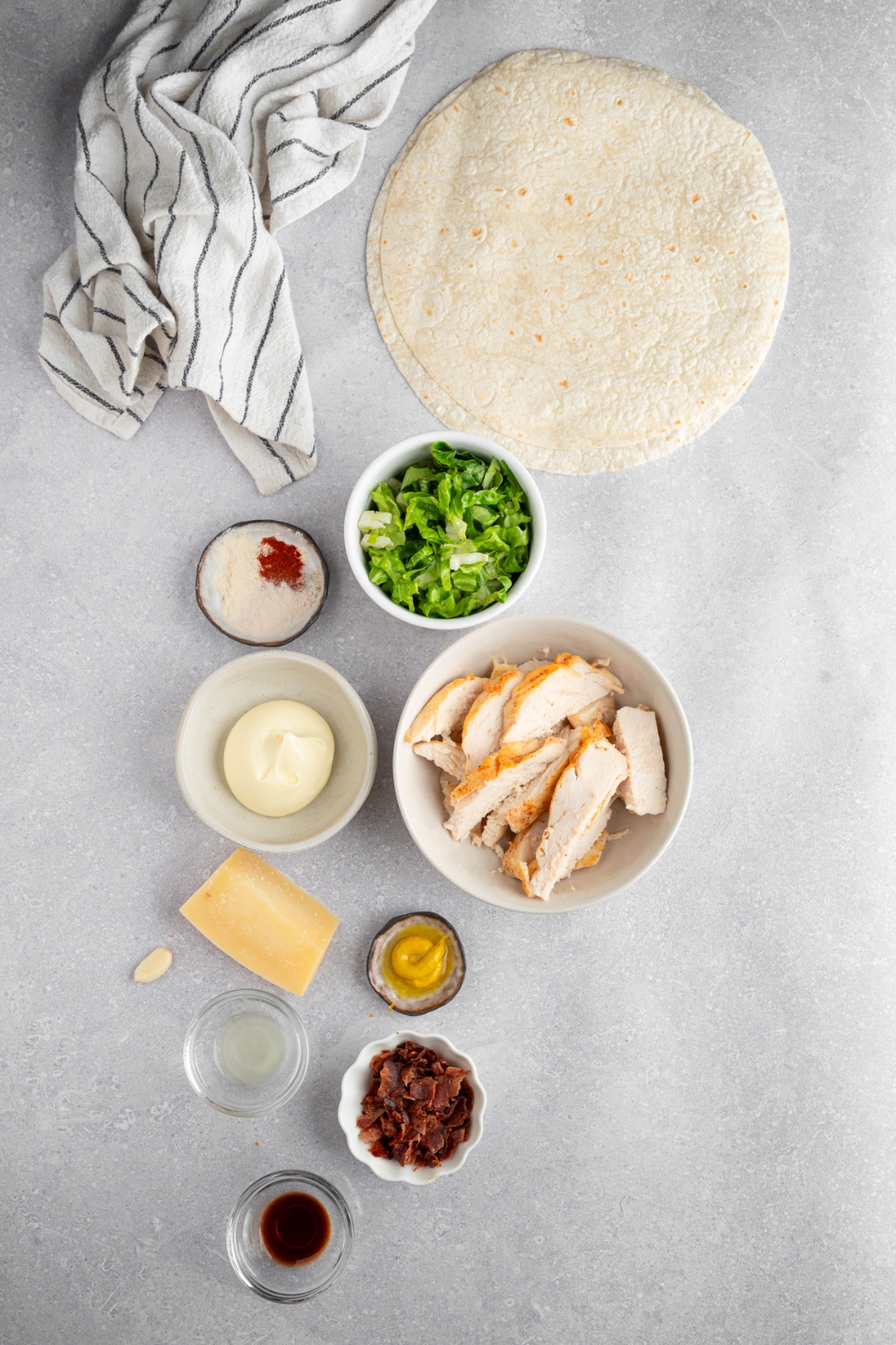 An overhead shot of several bowls in various sizes containing ingredients to make rotisserie chicken caesar wraps including tortillas, chicken, shredded lettuce, chopped bacon, mayo, mustard, and seasonings.