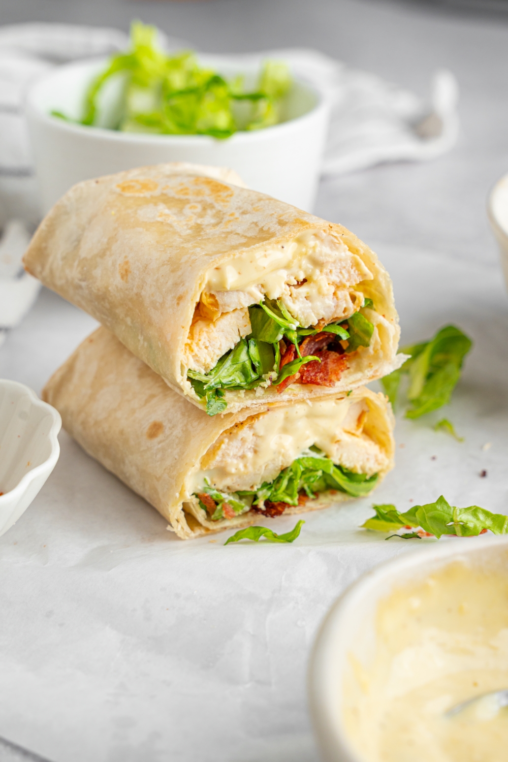 A rotisserie chicken caesar wrap sliced in half and stacked on one another. The wrap is on a white counter with a bowl of shredded lettuce.