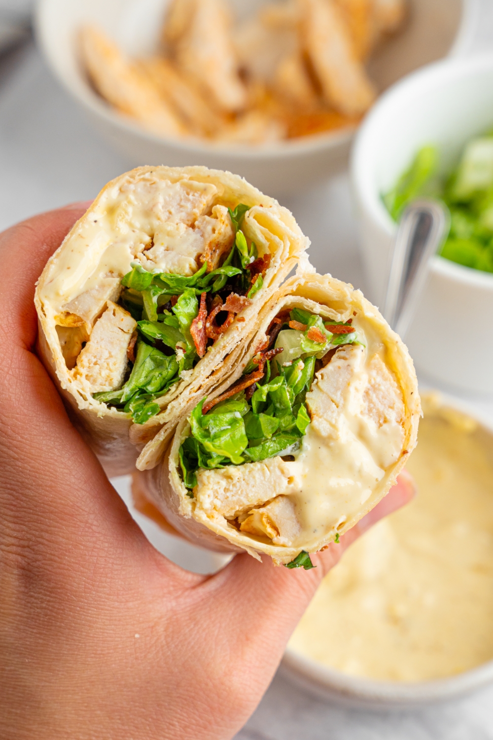 A hand holding a rotisserie chicken caesar wrap sliced in half. There is a bowl of caesar dressing, lettuce, and chicken blurred in the background.
