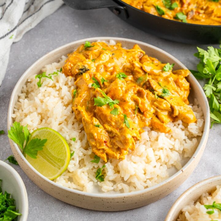 A white bowl with rotisserie chicken curry served with white rice and garnished with fresh parsley and lime. The bowl is on a white counter with a skillet of curry and white striped napkin.