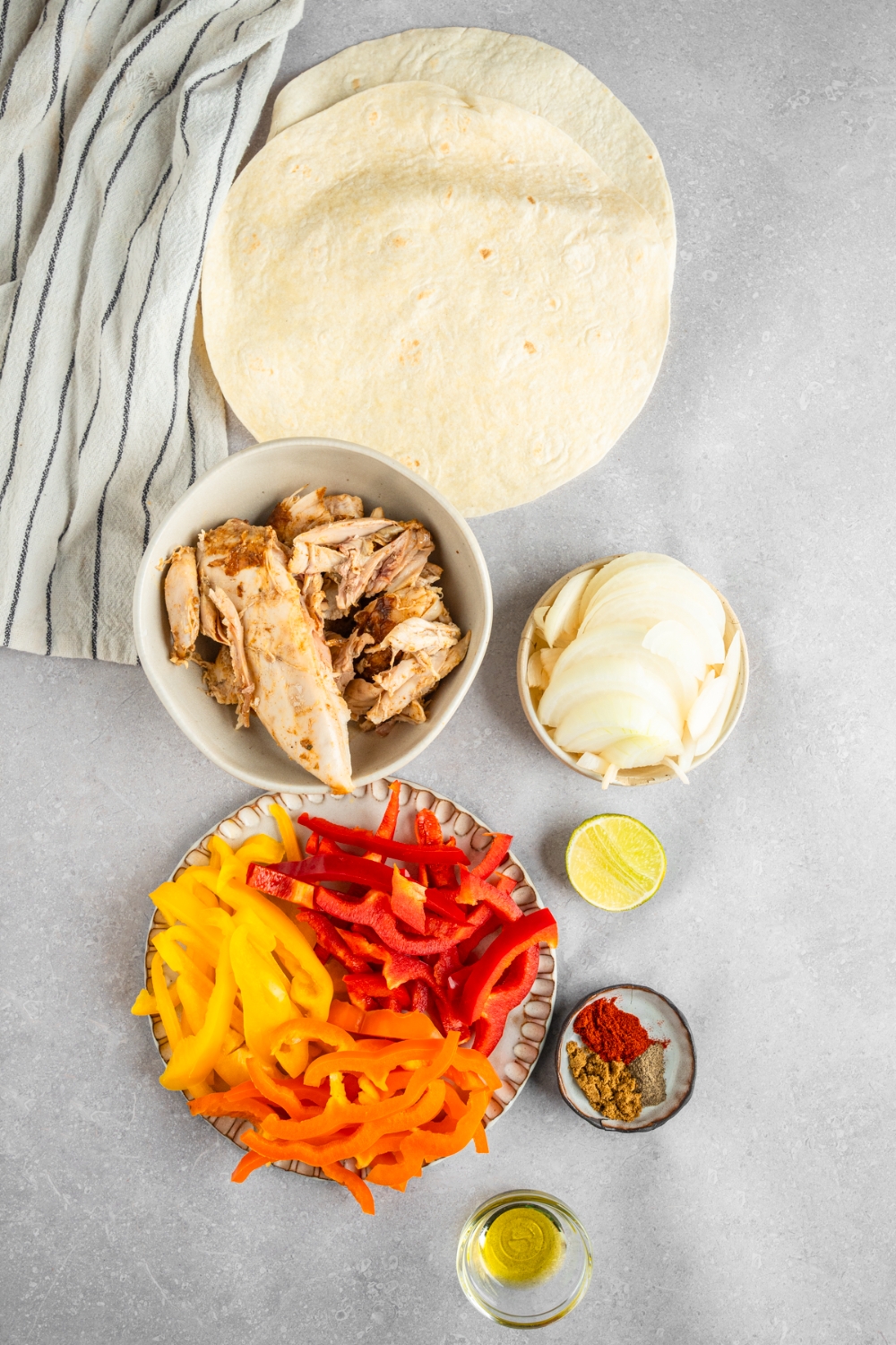 An overhead shot of several bowls in various sizes containing ingredients to make rotisserie chicken fajitas including pitas, shredded rotisserie chicken, sliced bell peppers, onion, lime, and seasonings.