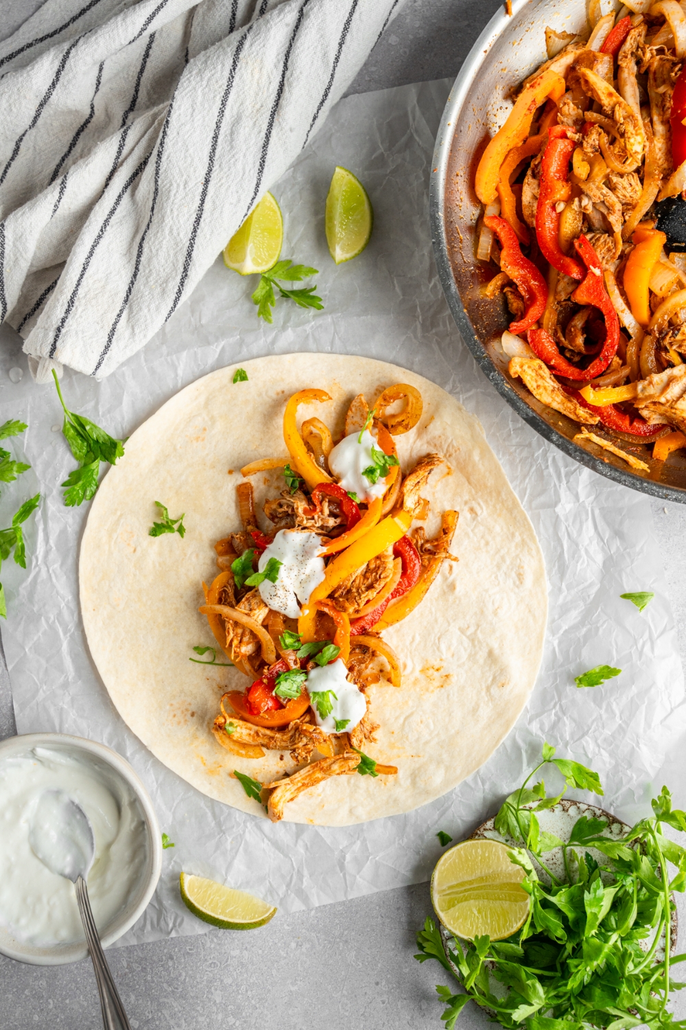 An open rotisserie chicken fajita topped with sour cream and fresh. cilantro. The fajita is on a piece of parchment paper on a white counter with a skillet of chicken fajitas and lime wedges.