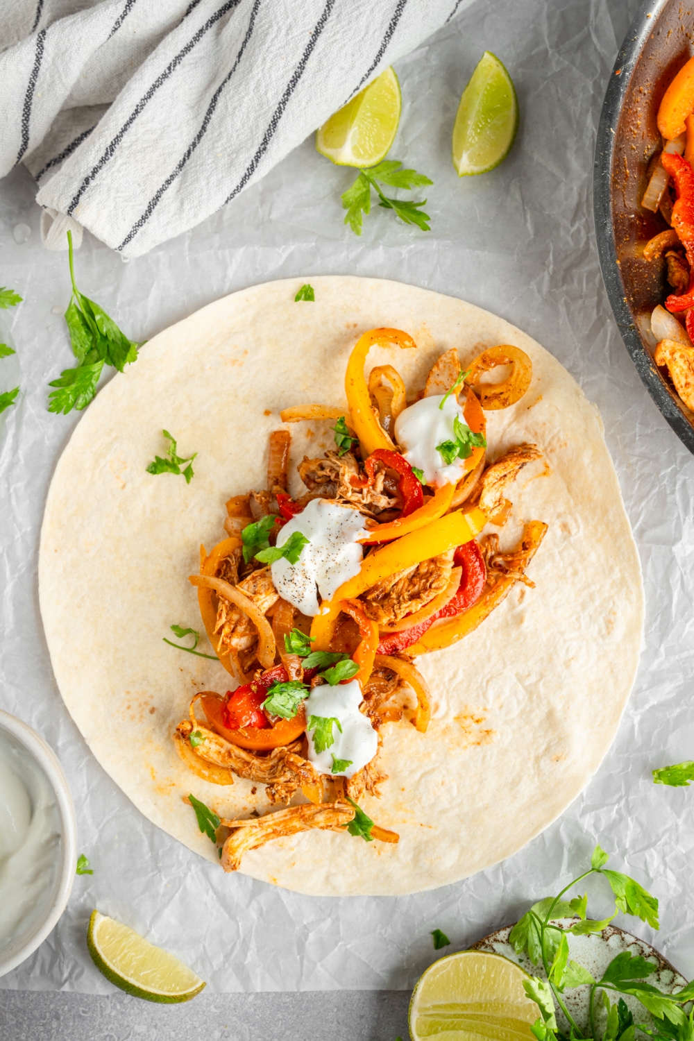 An open rotisserie chicken fajita topped with sour cream and fresh. cilantro. The fajita is on a piece of parchment paper on a white counter with a skillet of chicken fajitas and lime wedges.