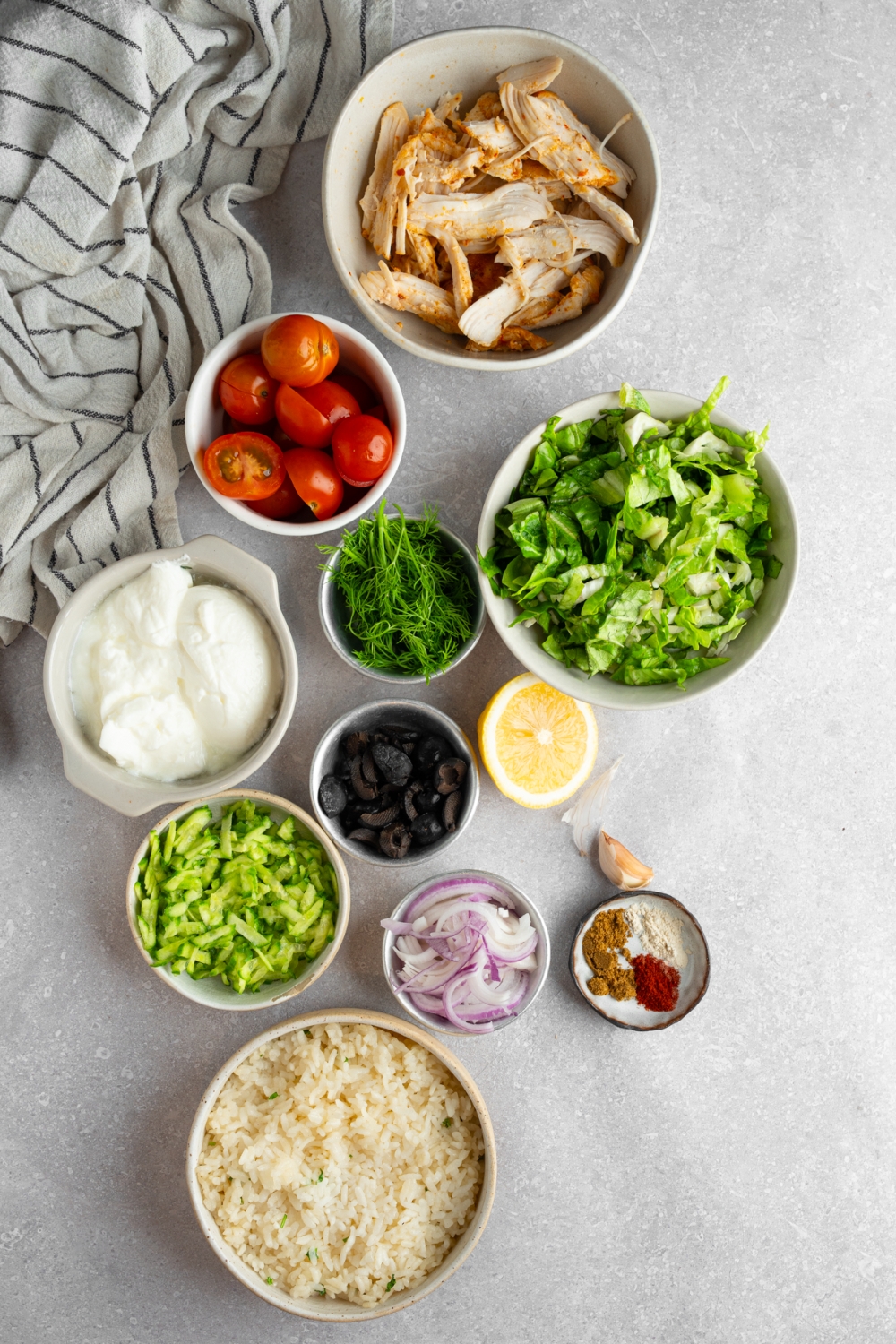 An overhead shot of several bowls in various sizes containing ingredients to make rotisserie chicken greek bowls including rotisserie chicken, tomatoes, shredded lettuce, dill, black olives, white rice, red onion, cucumber, greek yogurt, and seasonings.