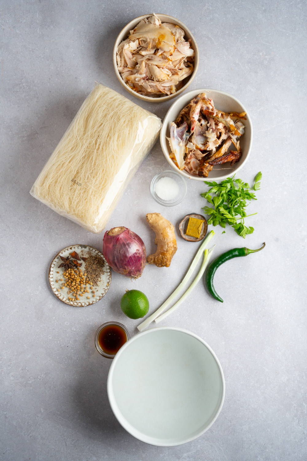 An overhead shot of several bowls in various sizes containing ingredients to make rotisserie chicken pho including uncooked pho noodles, shredded rotisserie chicken, red onion, ginger, fish sauce, water, jalapeno, basil, lime, and spices.