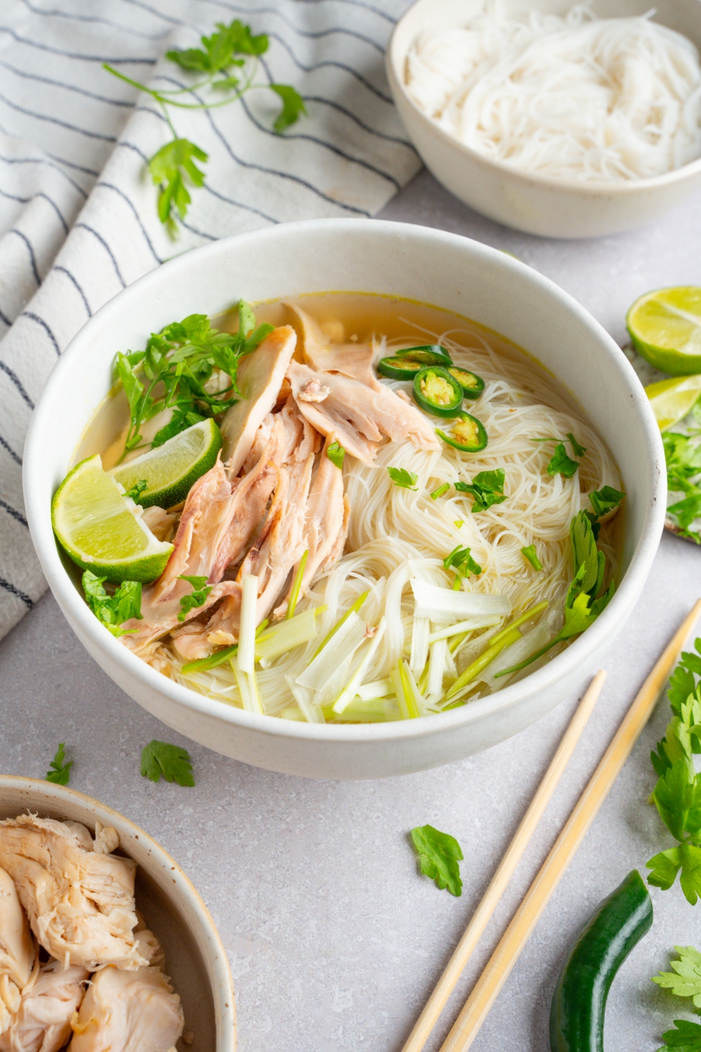 Rotisserie chicken pho served in a white bowl garnished with lime, fresh basil leaves, cilantro, bean sprouts, sliced jalapeno, and green onions. A pair of chopsticks is next to the bowl. The bowl is on a white counter with a white bowl of cooked pho noodles and a white striped napkin.