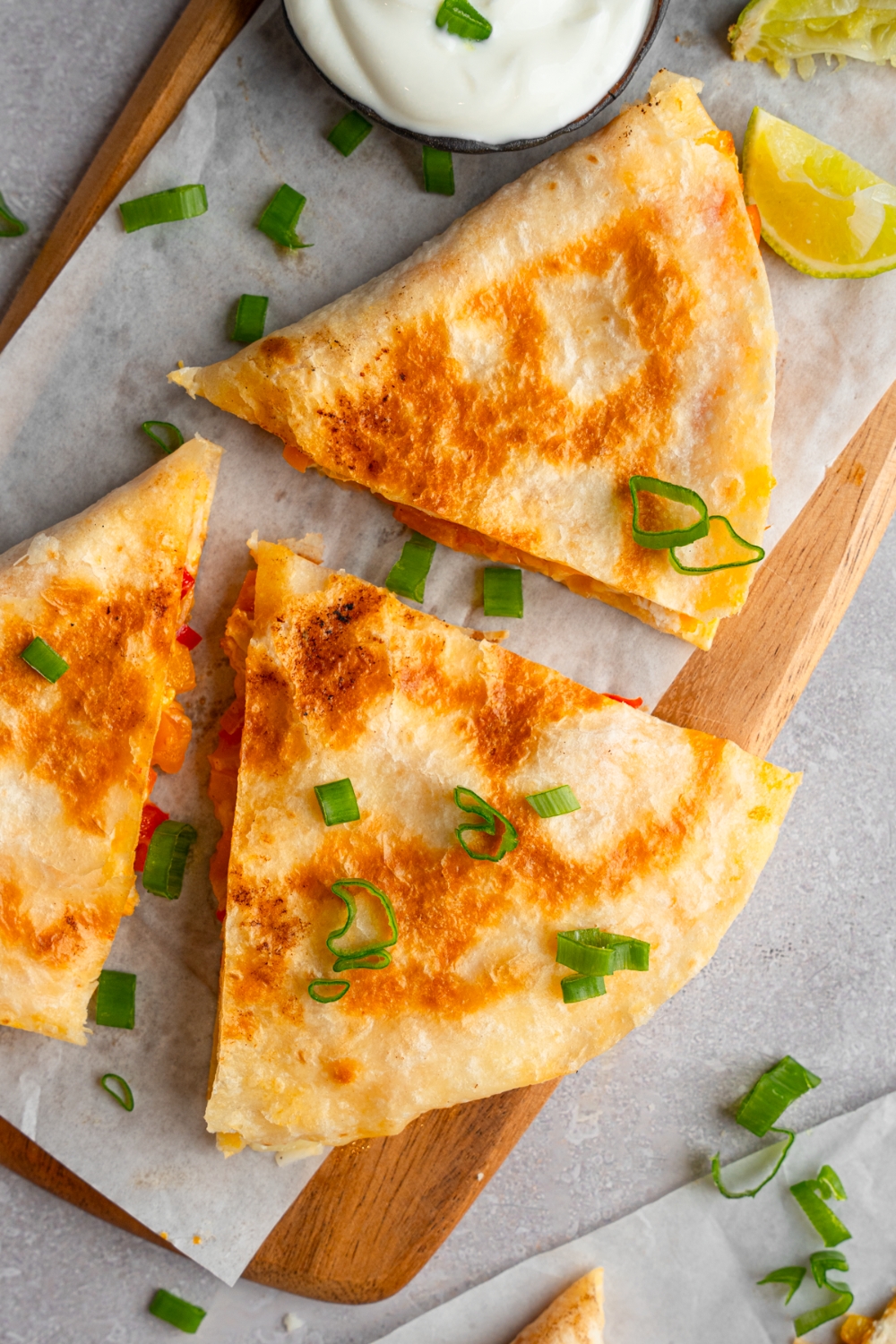 Three slices of rotisserie chicken quesadilla garnished with sliced green onion. The quesadilla is on a wooden board lined with parchment paper with a small cup of sour cream.