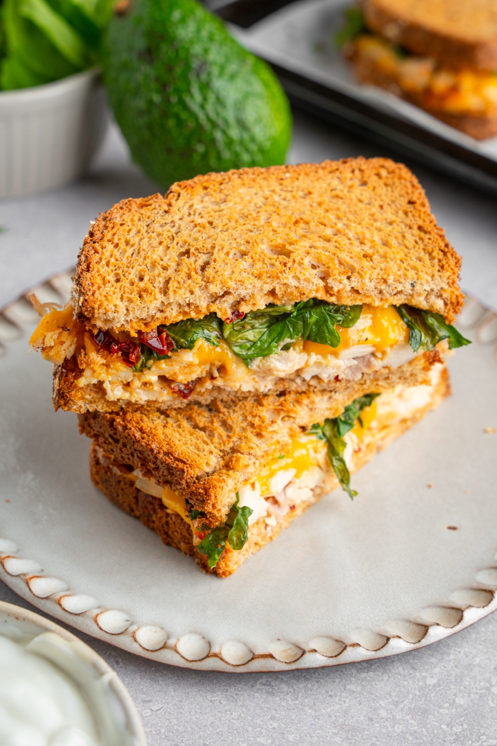 A rotisserie chicken sandwich with melted cheese, spinach, and chipotle mayo sliced in half and served on a ceramic plate. One half is stacked on top of the other. The plate is on a white counter with an avocado.