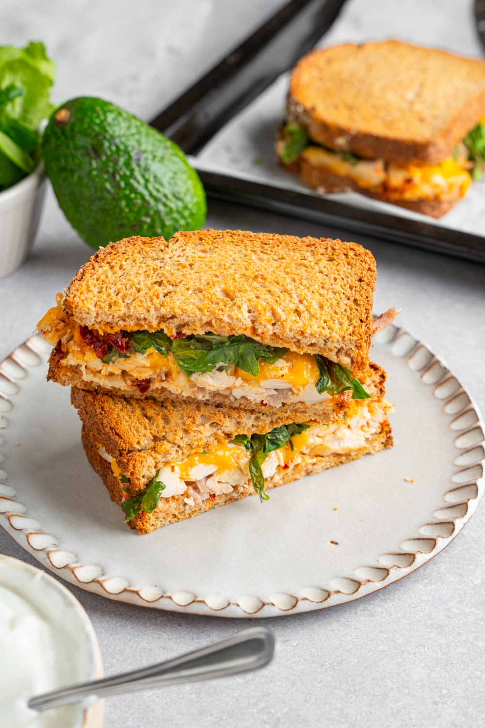 A rotisserie chicken sandwich with melted cheese, spinach, and chipotle mayo sliced in half and served on a ceramic plate. One half is stacked on top of the other. The plate is on a lined baking sheet with an additional sandwich and an avocado.