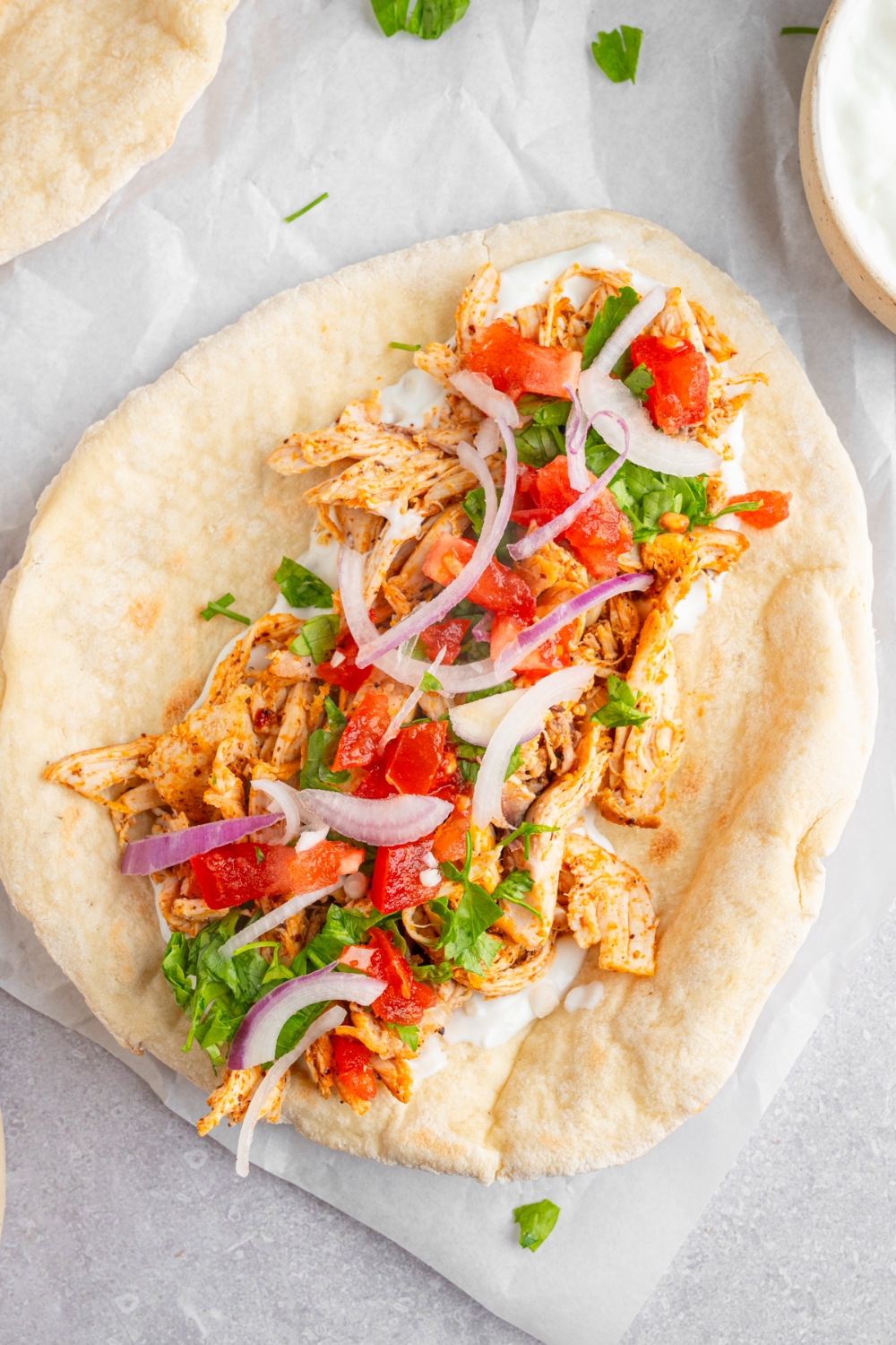 An open chicken shawarma including a pita topped with rotisserie chicken, garlic sauce, red onion, tomato, and fresh parsley. The parchment paper is on a white counter with additional pita and small bowl of garlic sauce.