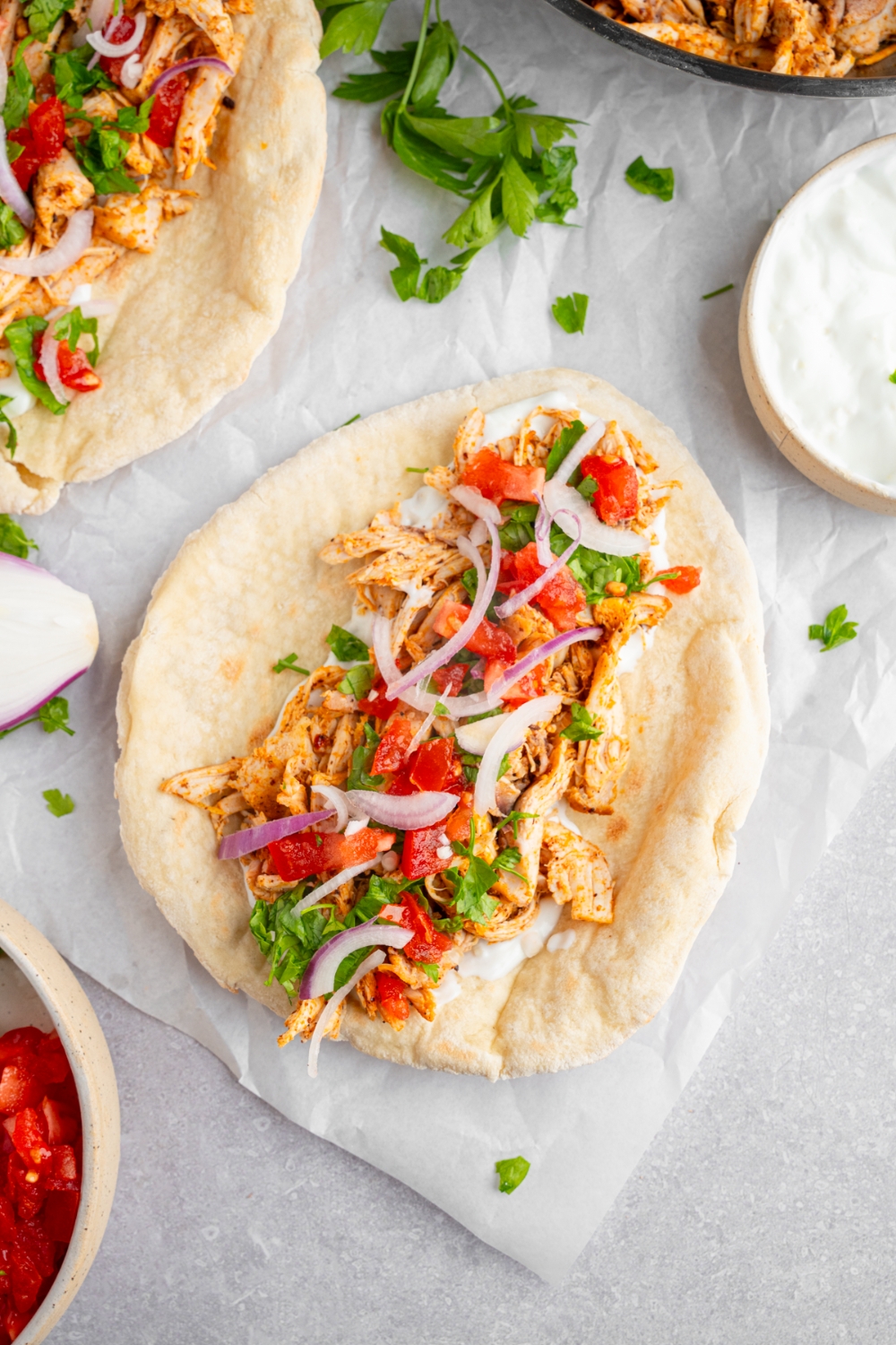 An open chicken shawarma including a pita topped with rotisserie chicken, garlic sauce, red onion, tomato, and fresh parsley. The parchment paper is on a white counter with additional pita and small bowl of garlic sauce.