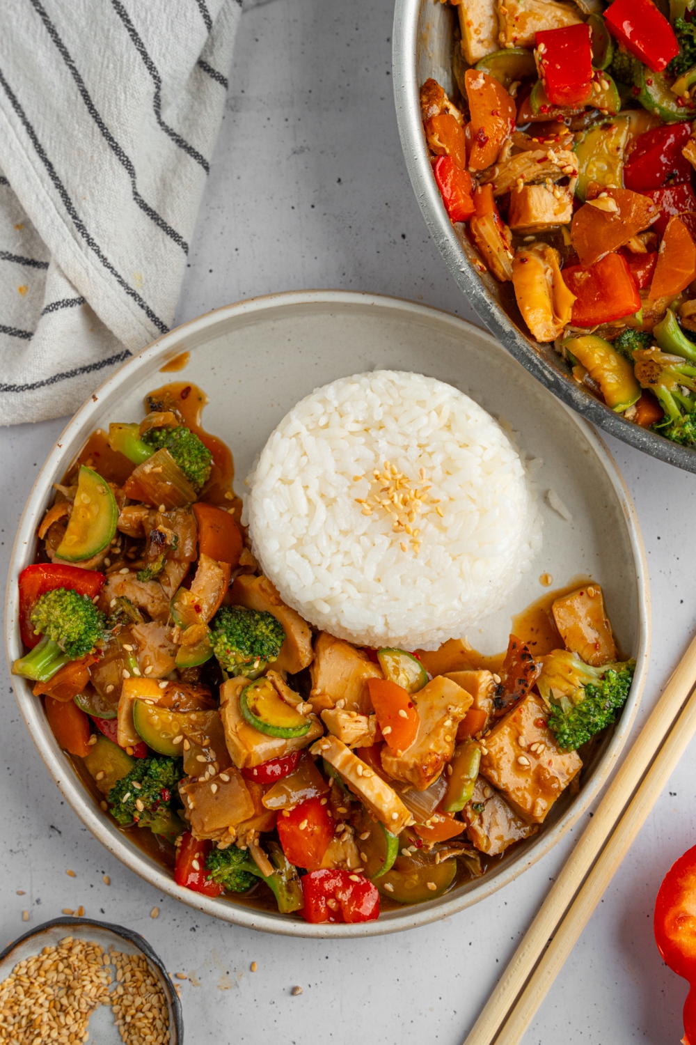 A plate of rotisserie chicken stir fry served with white rice. The plate is on a white counter with a white striped napkin and pair of chopsticks.