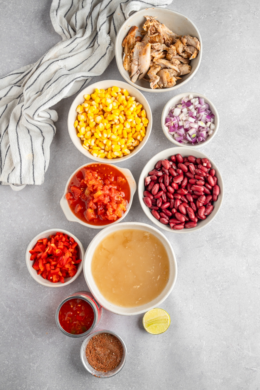 An overhead shot of several bowls in various sizes containing ingredients to make rotisserie chicken taco soup including rotisserie chicken, corn, red kidney beans, diced tomatoes, red onion, chicken broth, salsa, and seasonings.