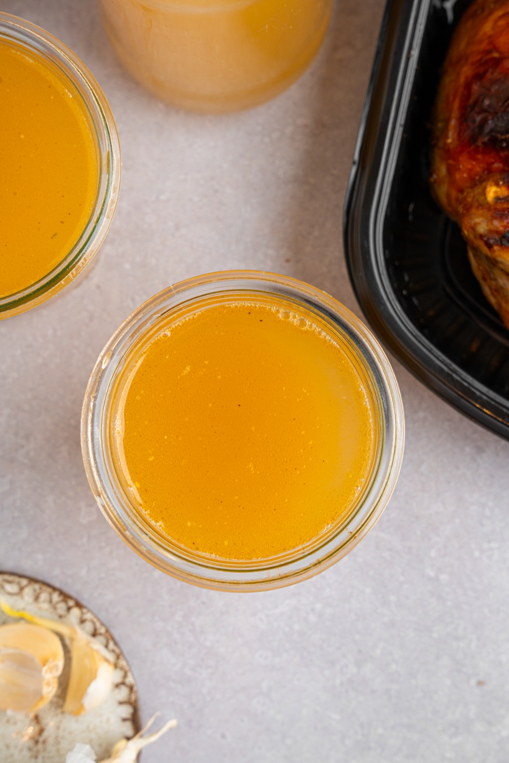 A glass jar with rotisserie chicken bone broth. The jar is on a white counter with a second jar and pitcher of bone broth. There is a tray of rotisserie chicken next to the jars.