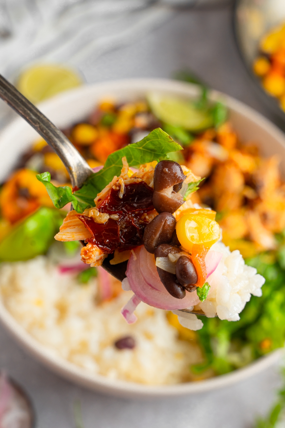 A close up of a fork with a bite of rotisserie chicken, rice, beans, and corn. There is a whole rotisserie chicken burrito bowl blurred in the background.