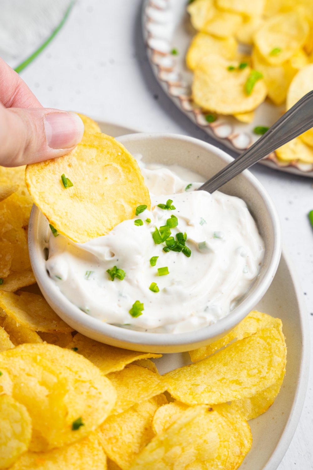 A hand dipping a potato chip into a bowl of sour cream and chive dip.