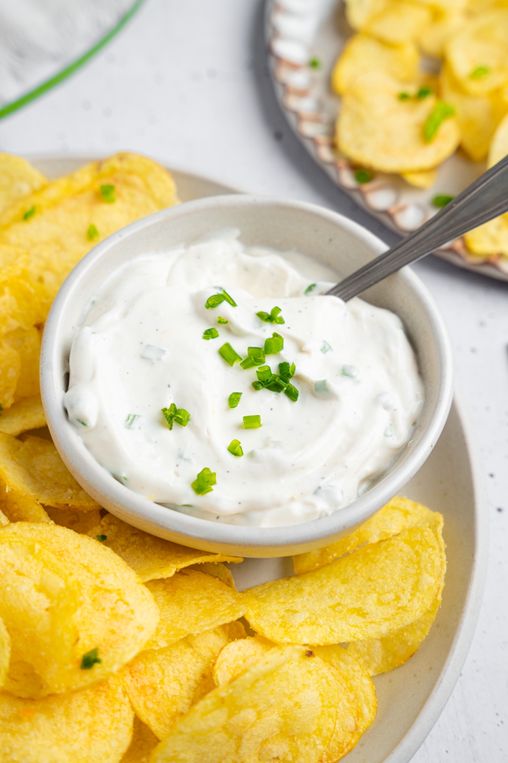 A bowl of sour cream and chive dip. I'm part of a white plate with potato chips.