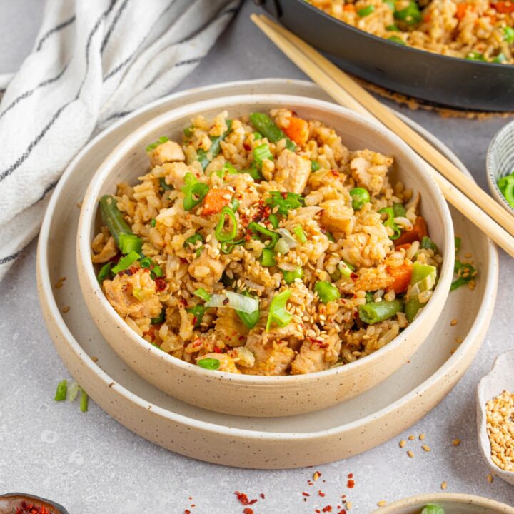 Fried rice with rotisserie chicken served in a ceramic bowl garnished with sliced green onion. The bowl is on a ceramic plate with a pair of chopsticks. The plate is on a white counter with a skillet of fried rice and bowls of garnishes.