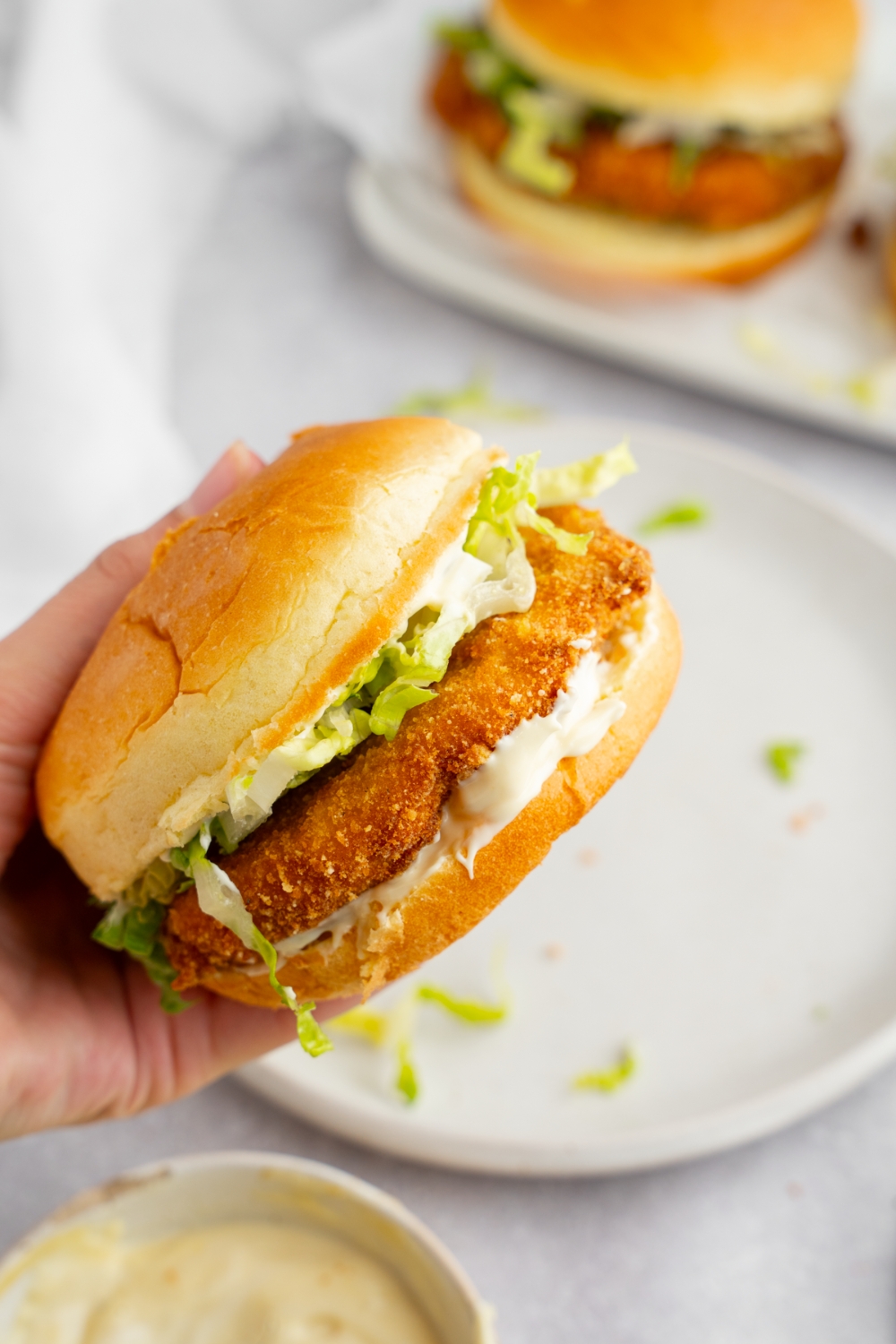 A hand holding a homemade McDonald's chicken sandwich. There is a white and extra plate with a chicken sandwich blurred in the background.