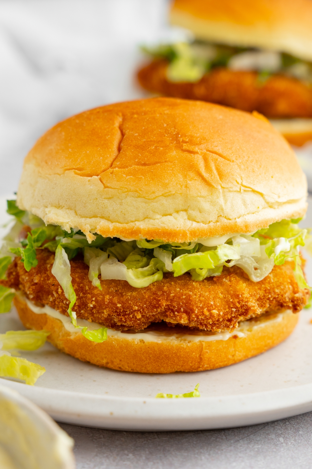 A homemade McDonald's chicken sandwich with shredded lettuce on a white plate. The plate is on a white counter with an additional sandwich.