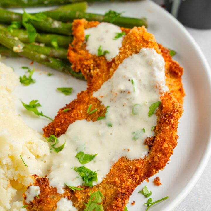 A white ceramic plate with homemade Cheesecake factory chicken costoletta with a lemon cream sauce. The chicken is served with asparagus and mashed potatoes garnished with fresh parsley.