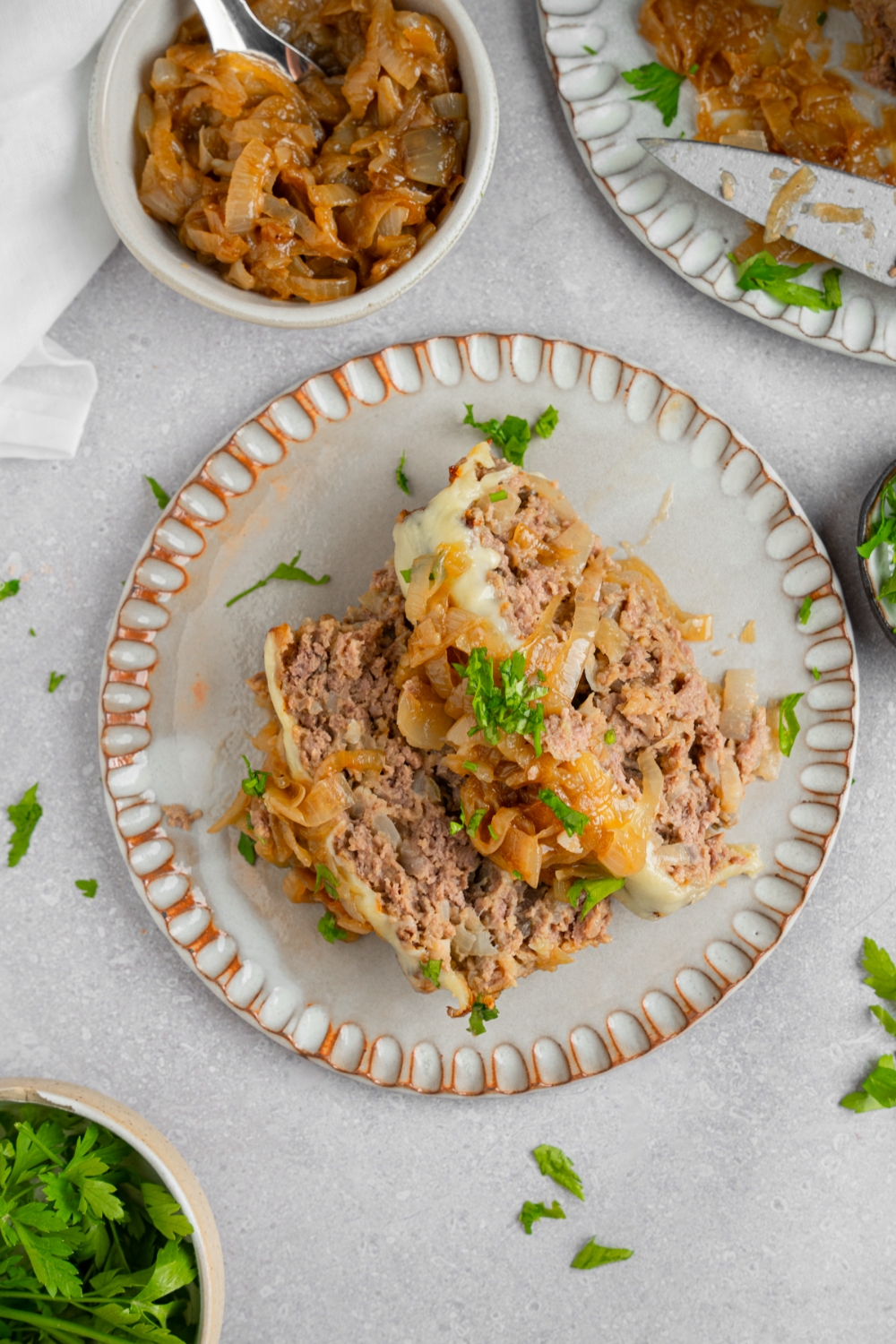 A ceramic plate with two slices of french onion meatloaf garnished with fresh parsley. The plate is on a white counter with a small bowl of carmelized onions.