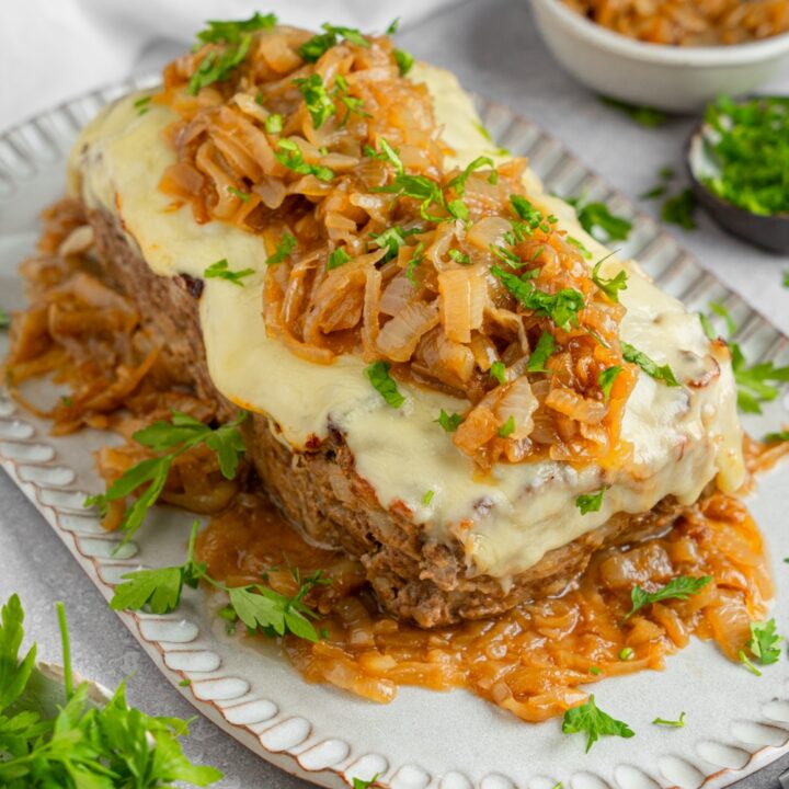 French onion meatloaf on a ceramic plate. The meatloaf is topped with carmelized onions and garnished with fresh parsley. The plate is on a white counter with a bowl of carmelized onions.