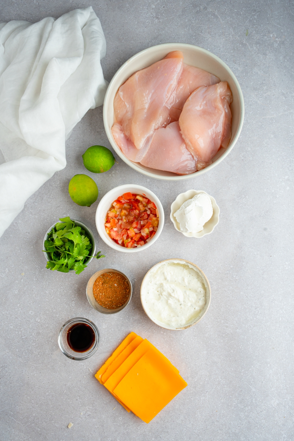An overhead shot of several bowls in various sizes containing ingredients to make homemade Applebee's fiesta lime chicken including chicken breasts, lime, salsa, sour cream, slice cheese, and seasonings.