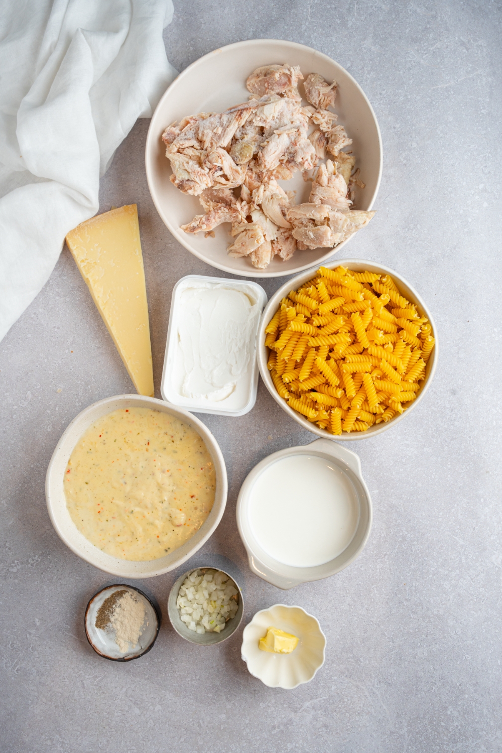 An overhead shot of several bowls in various sizes containing ingredients to make homemade Buffalo Wild Wings garlic parmesan chicken pasta including pasta, shredded chicken, cream cheese, Buffalo Wild Wings garlic parmesan sauce, heavy cream, and seasonings.
