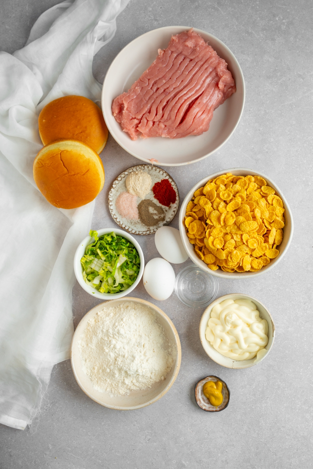An overhead shot of several bowls in various sizes containing ingredients to make homemade McDonald's chicken sandwich including ground chicken, two buns, cornflakes, shredded lettuce, flour, egg, mayo, and seasonings.