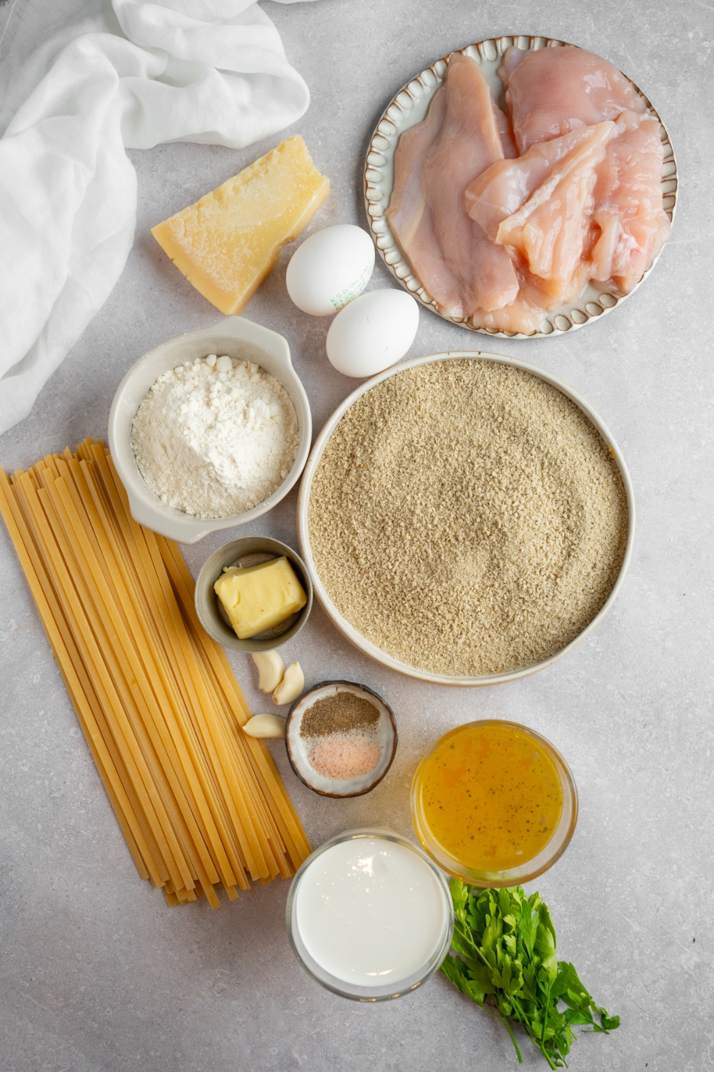 An overhead shot of several bowls in various sizes containing ingredients to make homemade Olive Garden chicken fritta including chicken breasts, fettuccine pasta, cheese, breadcrumbs, cream, chicken stock, butter, eggs, fresh parsley, and seasonings.