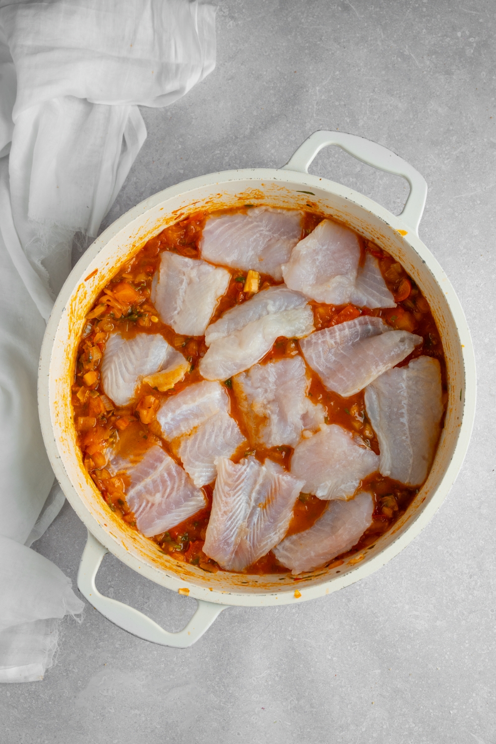 A stock pot with uncooked fish on top of a stew base. The pot is on a white counter with a white napkin.