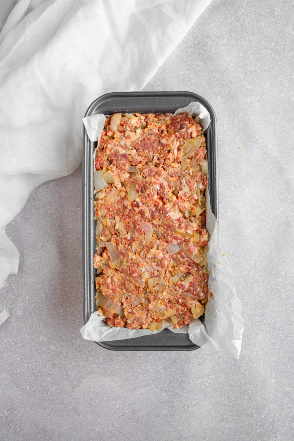 A loaf pan lined with parchment paper with uncooked french onion meatloaf mixture. The pan is on a white counter with a white napkin.