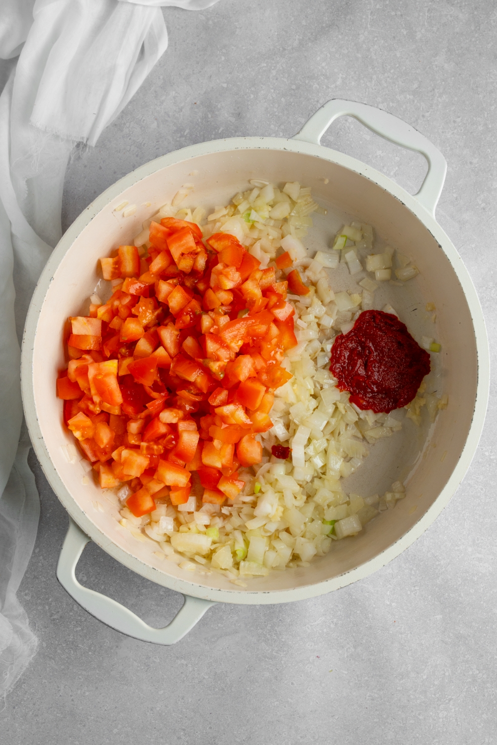 A stock pot with diced onion, tomatoes, and tomato paste. The pot is on a white counter with a white napkin.