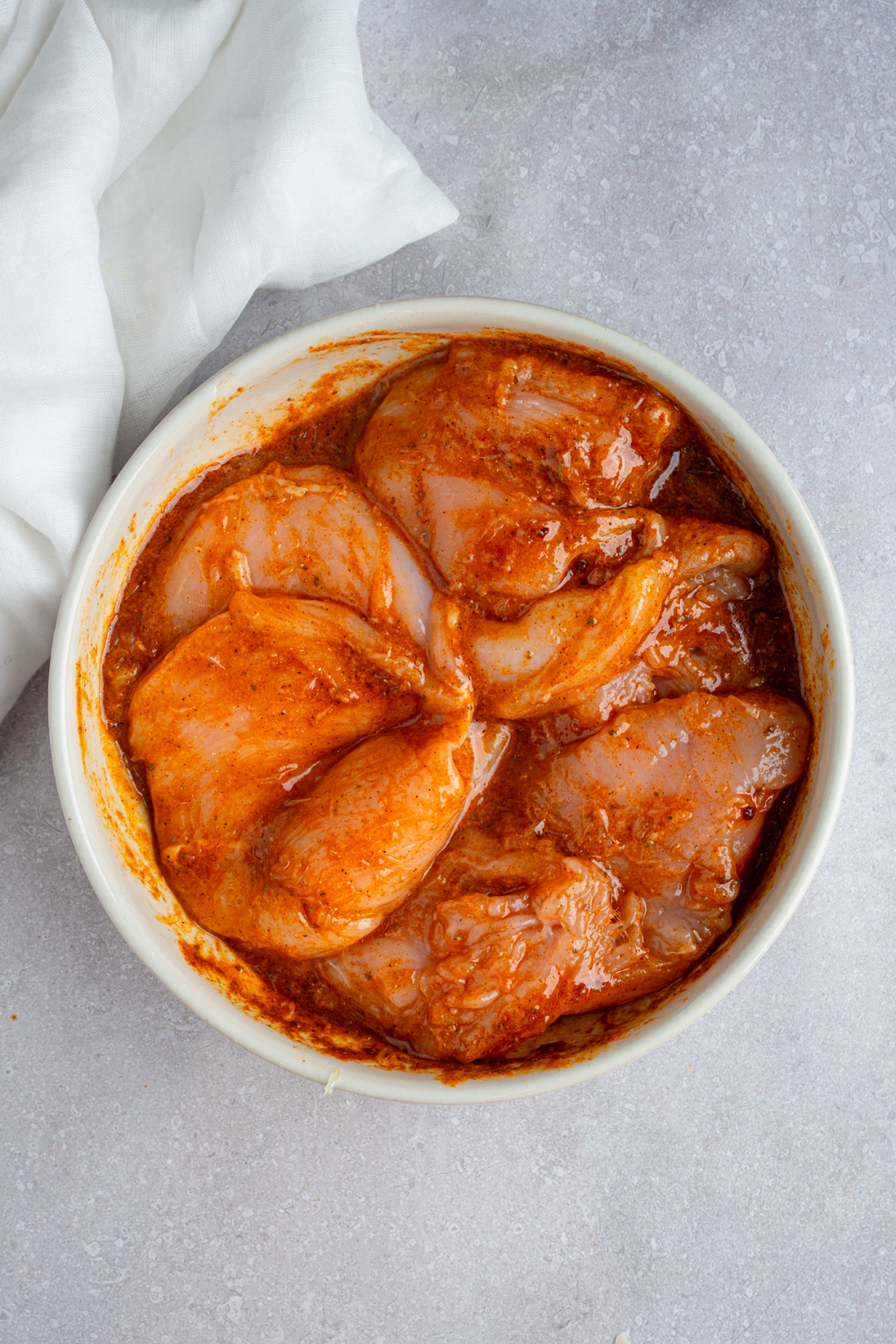 A white bowl with four uncooked chicken breasts marinating in a fiesta lime marinade. The bowl is on a white counter with a white cloth napkin.