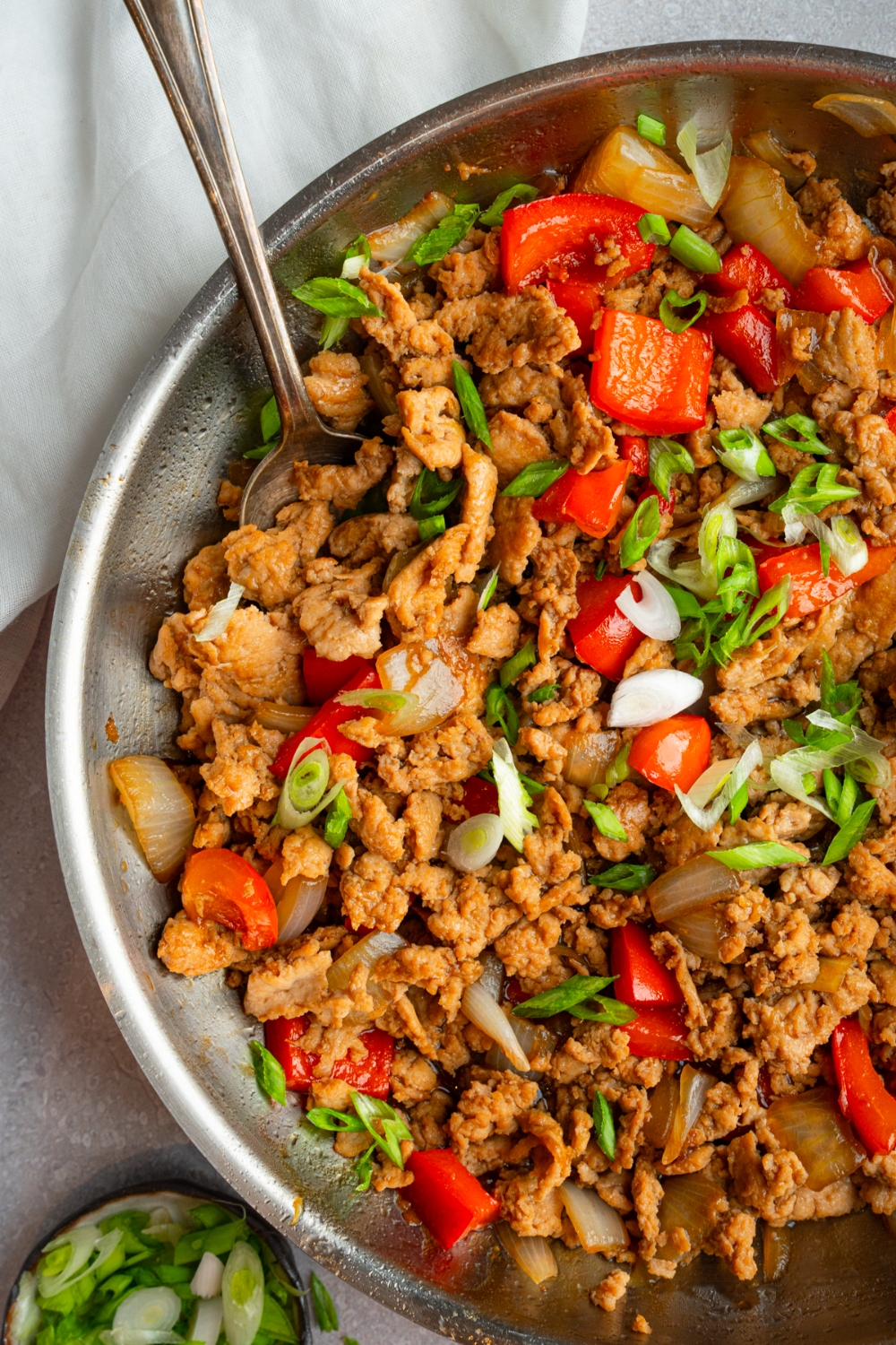 A skillet with minced chicken stir fry garnished with sliced green onions and served with a spoon. The skillet is on a white counter with a small plate of garnishes.