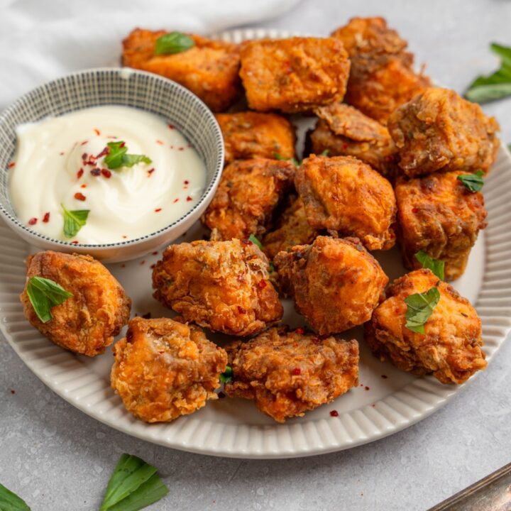 A plate of fried salmon nuggets garnished with fresh basil and served with a small ramekin of dipping sauce. The plate is on a white counter with a white cloth napkin.